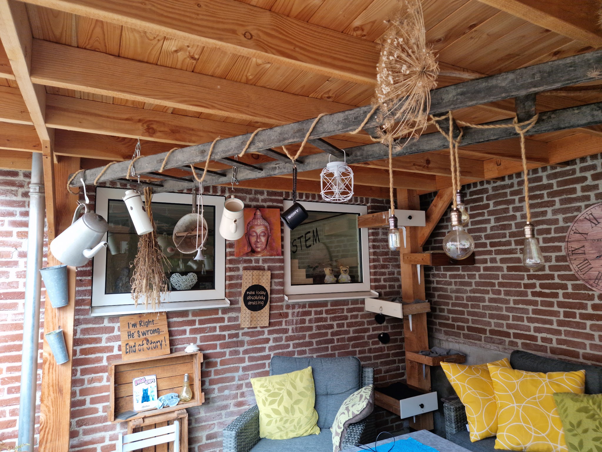 Onder het afdak: houten balken tegen een bakstenen muur, loungebank met gele kussens