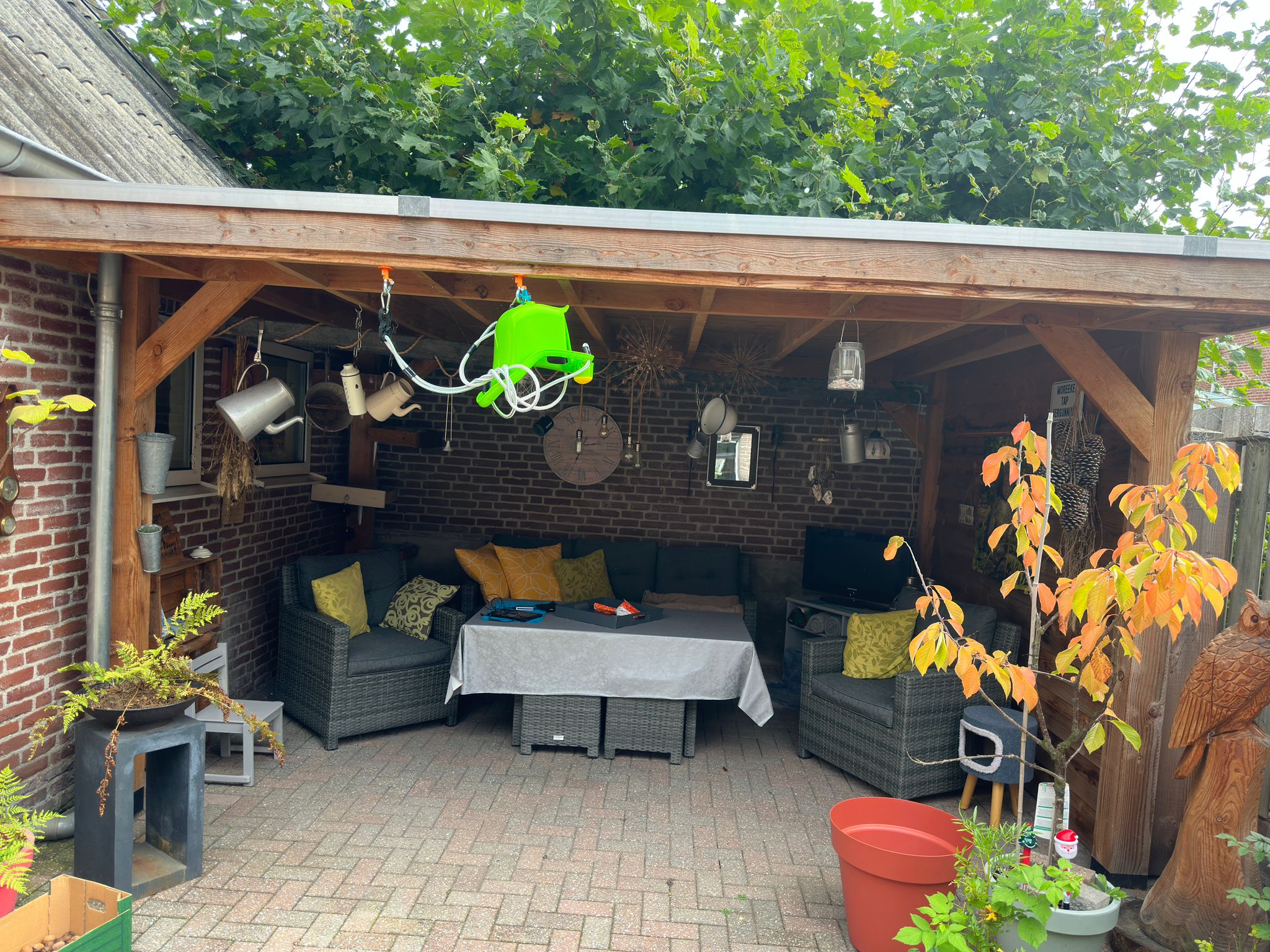 Houten afdak in de zomer boven een terras met loungeset en planten, boom erachter